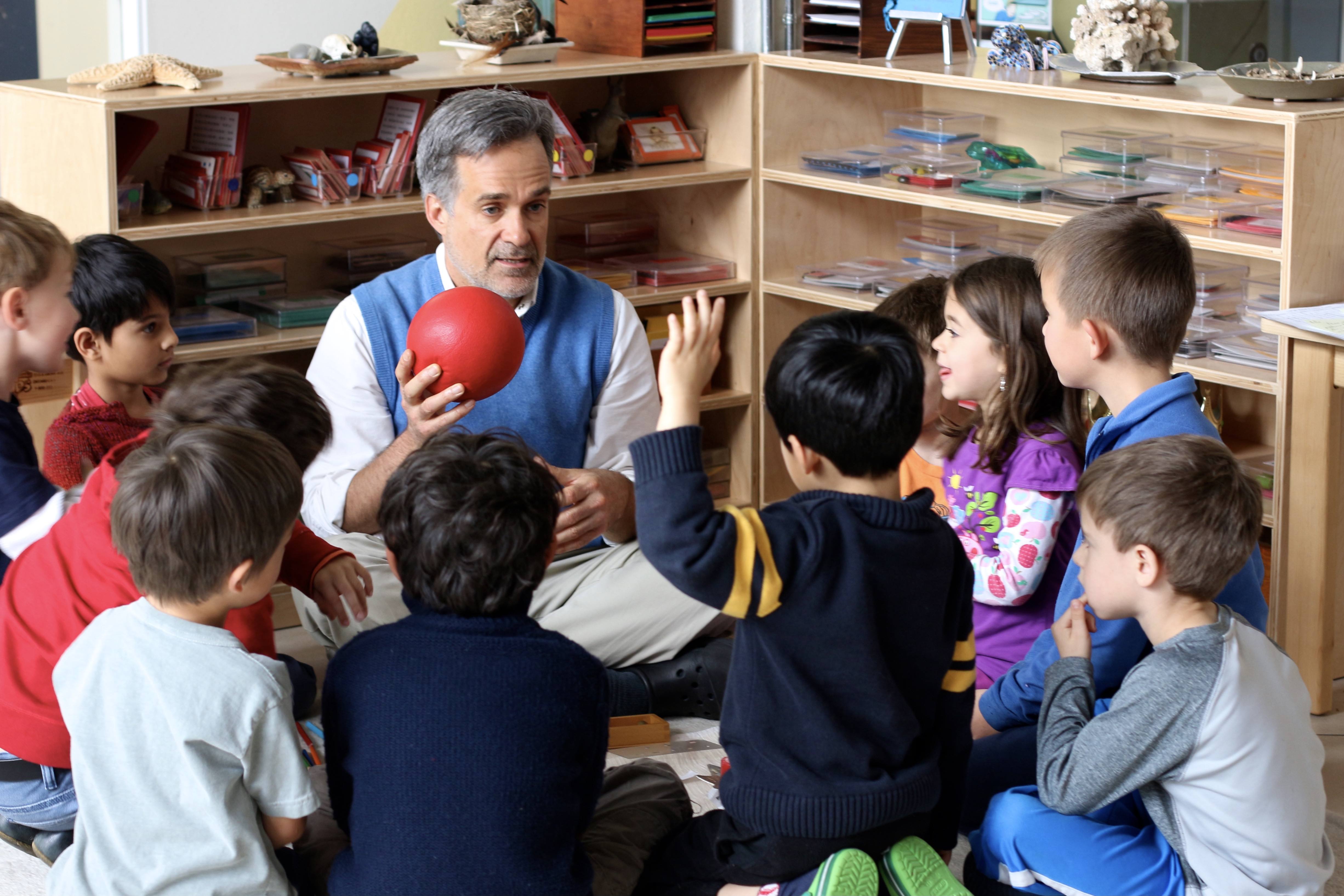 An educator sitting with children in a Montessori classroom, sharing a lesson with a red sphere